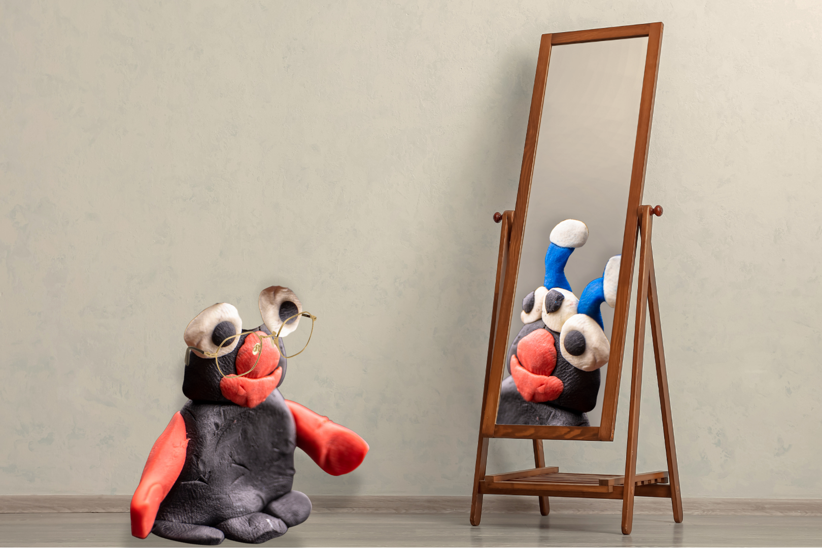 Black clay figure with orange hands, nose, and mouth wearing glasses and looking into a mirror, with a reflected clay figure that has three eyes and antenna‑like features.