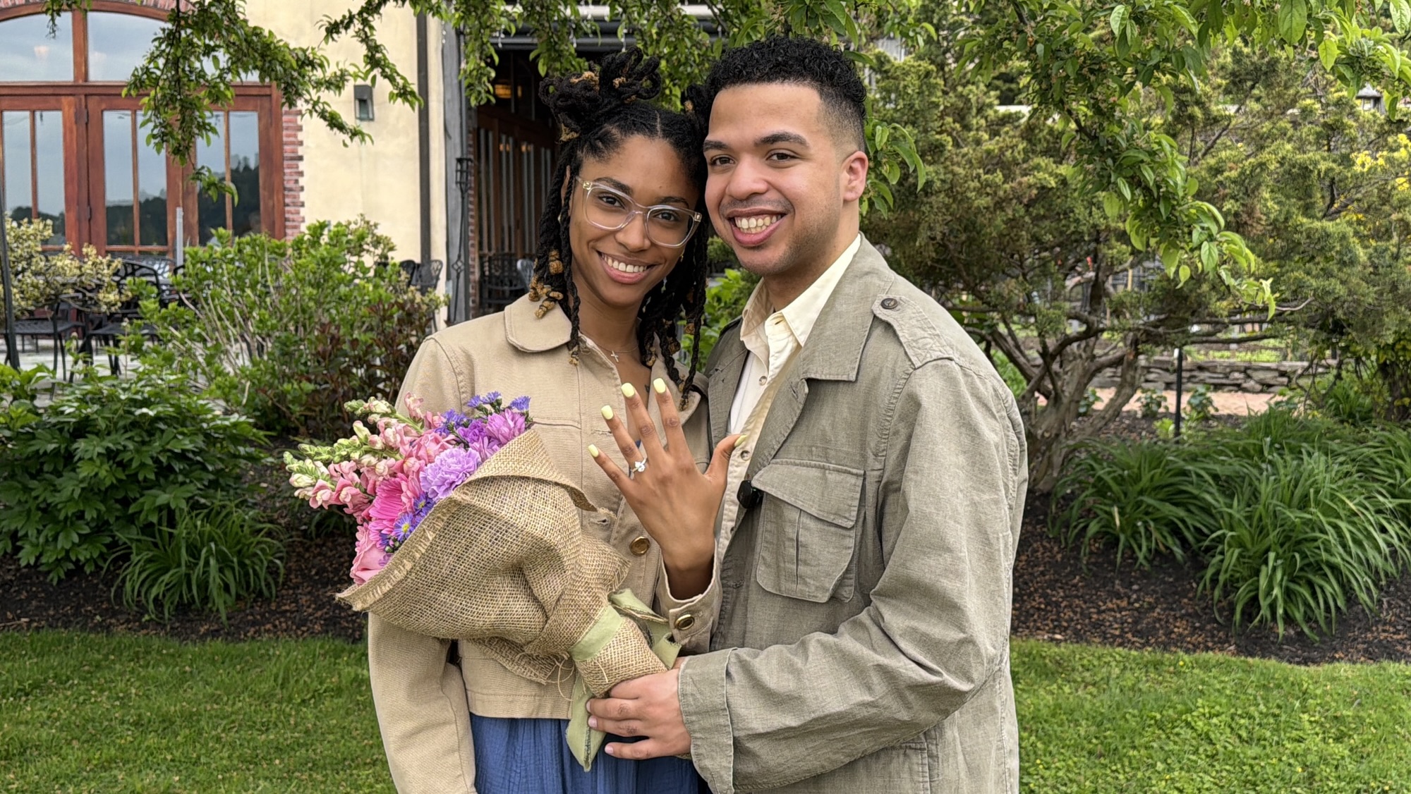 Justin Genao and Daina Jameison smiling outside Harvest on Hudson surrounded by greenery, with Justin holding a flower bouquet and Daina showing her engagement ring